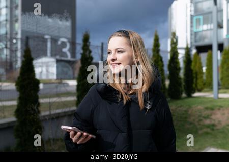 Jeune femme sourit tout en utilisant un smartphone à l'extérieur dans un cadre urbain moderne au cours d'un après-midi nuageux Banque D'Images