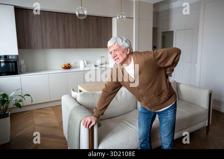 Douleurs dorsales. Homme âgé malheureux penché dans des poignées de douleur blessé dans le bas du dos, debout près du canapé de la maison Banque D'Images