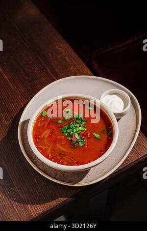Un bol de soupe de tomate rouge vif est placé sur une table en bois rustique. La soupe est garnie d'herbes vertes fraîches et accompagnée d'un petit côté de Banque D'Images