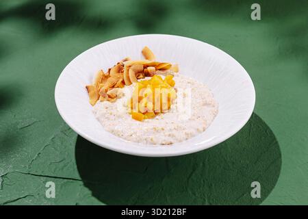 Délicieux bol de flocons d'avoine garni de mangue fraîche et de noix de coco grillée sur une surface verte vibrante Banque D'Images