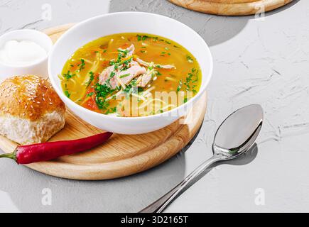 Un bol copieux de soupe de nouilles au poulet est assis sur un plateau en bois, accompagné d'un rouleau de graines de sésame, une cuillère de crème sure, et un piment rouge, prêt t Banque D'Images