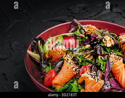 Salade de saumon frais composée de légumes verts mélangés, tomates cerises et graines de sésame servie dans un bol rouge sur fond de texture foncée Banque D'Images