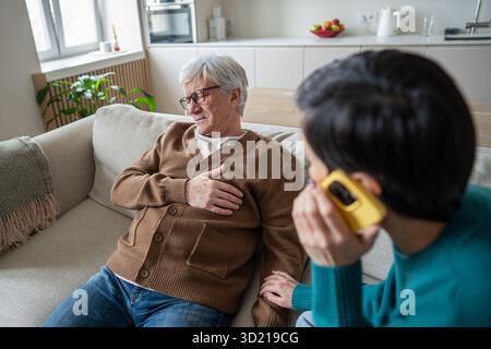 Homme âgé serrant la poitrine tout en étant assis sur la chaise, la femme appelle les services d'urgence avec le téléphone à proximité Banque D'Images