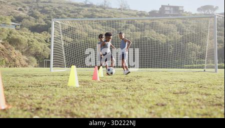 Trois garçons pratiquant des exercices de football dans un terrain herbeux, avec balle et cônes près du but Banque D'Images