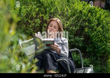 Détendu femme heureuse lisant le livre dans la chaise sur la pelouse dans le parc de jardin. Loisirs lecture habitude dans la nature Banque D'Images