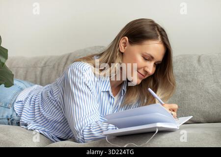 Femme souriante écrit des notes dans un journal intime couché sur un canapé confortable Banque D'Images