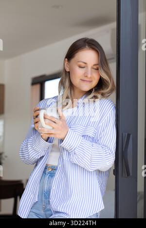 Femme positive en chemise rayée tient une tasse à café sur la terrasse Banque D'Images