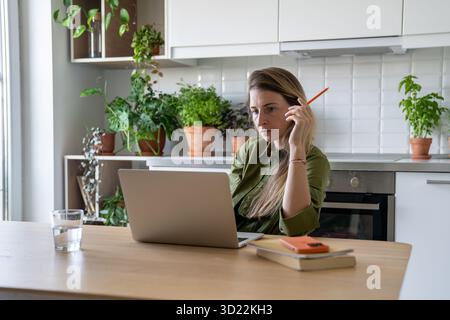Femme fatiguée réfléchie travaillant sur ordinateur portable, classes virtuelles en ligne entourées de livres et de verre d'eau Banque D'Images