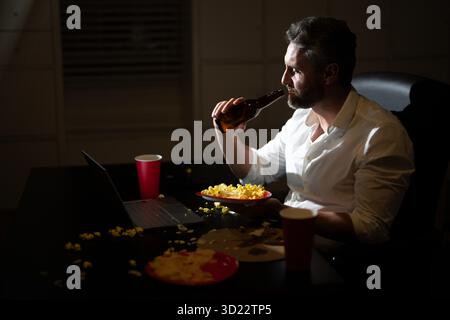 Homme d'affaires manger de la pizza en heures supplémentaires. Homme occupé des années 50 mangeant le dîner et travaillant en ligne au bureau de nuit. Entrepreneur travaille tard dans la nuit tout en profitant d'un Banque D'Images