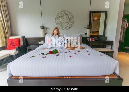 Femme dans la chambre d'hôtel se relaxant sur le lit avec des pétales de rose, draps blancs. Banque D'Images