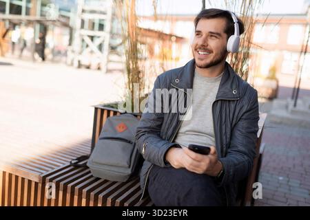 Jeune homme dans des écouteurs avec un sac à dos s'éloigne du temps en écoutant de la musique dans des écouteurs dans la rue Banque D'Images
