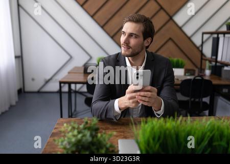 Un homme d'affaires en vêtements d'affaires avec un téléphone portable dans les mains est assis dans le bureau et regarde par la fenêtre Banque D'Images