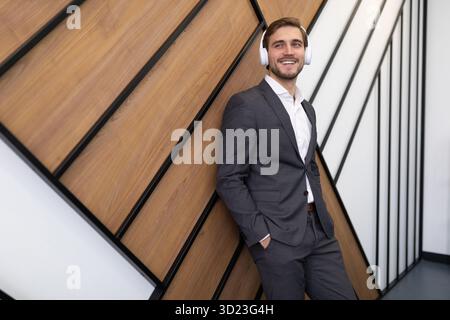 Jeune homme d'affaires dans des écouteurs dans un costume d'affaires écoutant de la musique appuyée contre le mur dans le bureau Banque D'Images