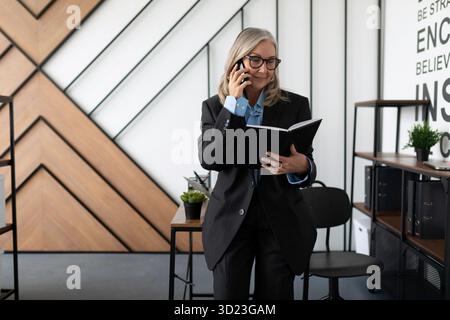 Femme gestionnaire de 50 ans dans un bureau stérile parle sur un téléphone portable avec un journal dans ses mains Banque D'Images