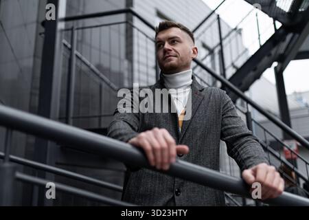 Portrait brutal d'un homme d'âge mûr sur un escalier en métal gris près d'un bâtiment moderne Banque D'Images