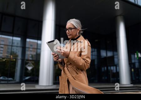 Femme adulte senior d'affaires avec une tablette marchant dans la rue à côté de l'immeuble de bureaux, femme européenne de 50 ans, Busin Banque D'Images