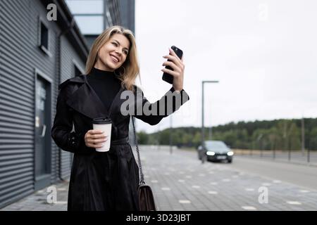 Femme d'affaires moderne d'âge moyen réussie parlant sur l'appel vidéo avec une tasse de café sur le fond d'un buil de bureau Banque D'Images