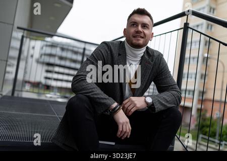 Élégant homme caucasien assis sur les marches souriant regardant la caméra Banque D'Images