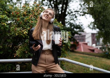 Jeune femme avec une tasse de café par téléphone portable écoutant de la musique sur des écouteurs tout en se relaxant dans le parc Banque D'Images