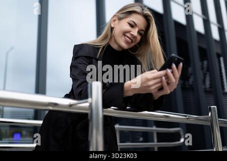 Femme d'affaires élégante sur le fond d'une façade en verre avec un téléphone dans ses mains souriant Banque D'Images