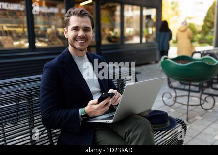 Un jeune homme avec un ordinateur portable et un téléphone est assis sur un banc à côté d'un café de la ville dans la rue, les principes et les règles du busi réussi Banque D'Images