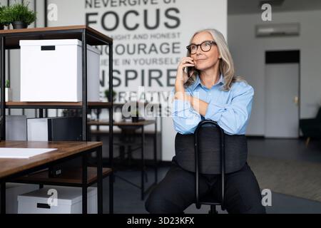 Top Manager femme d'affaires plus âgée d'âge moyen s'assoit sur une chaise et parle sur un téléphone portable, travail avec le concept d'objections Banque D'Images