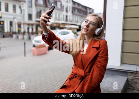Élégante jeune femme caucasienne dans des écouteurs fait selfie dans la ville Banque D'Images