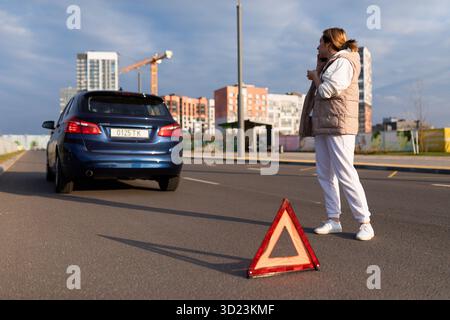 Une conductrice sur une autoroute à côté d'une voiture en panne parlant sur un téléphone portable à côté d'un triangle d'arrêt d'urgence, coffre-fort routier Banque D'Images