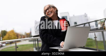 Femme d'affaires âgée élégante travaille sur un ordinateur portable en ligne sur la terrasse d'un immeuble de bureaux et parle sur un téléphone portable pendant d Banque D'Images