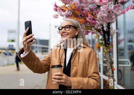 Femme âgée parlant sur un appel vidéo en utilisant le téléphone à l'entrée du magasin à l'extérieur Banque D'Images