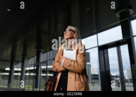 Une femme d'affaires de cinquante ans en âge de retraite quitte l'immeuble de bureaux regarde autour de lui avec un ordinateur portable dans ses mains Banque D'Images