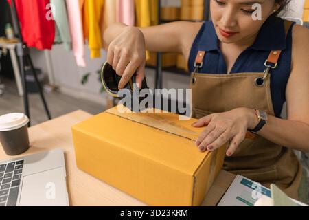 Jeune femme entrepreneure scellant soigneusement le paquet avec du ruban adhésif, préparant les commandes de marchandises, gérant l'expédition et la livraison pour son succès en ligne Banque D'Images