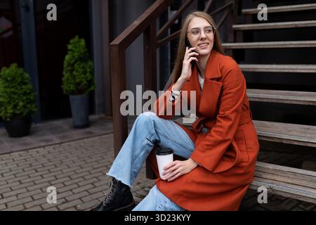 Portrait d'une belle jeune femme élégante dans un manteau orange assis sur les escaliers et parlant sur un téléphone portable, concept de yo Banque D'Images