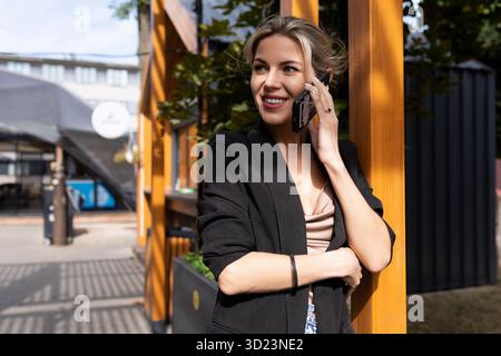 Femme d'âge moyen parlant au téléphone sur le fond de la ville regarde loin avec un sourire sur son visage Banque D'Images