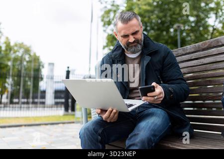 L'homme mature avec la barbe et les cheveux gris travaille à l'extérieur avec ordinateur portable et téléphone Banque D'Images