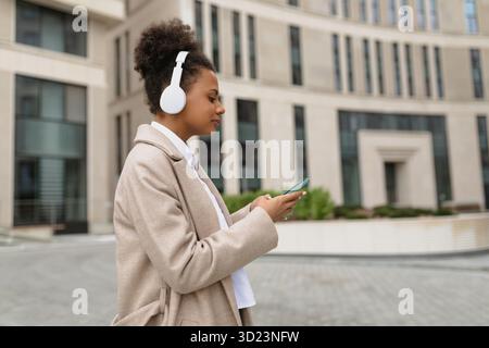 Jeune femme afro-américaine se dépêchant de travailler en écoutant de la musique dans des écouteurs via un téléphone portable dans le contexte d'un offic Banque D'Images