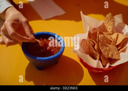 Tremper à la main les chips de maïs dans un bol de salsa sur une table orange vif à côté du panier de chips. Californie, États-Unis Banque D'Images