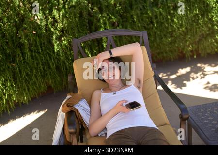 Jeune femme adulte se relaxant sur une chaise longue à l'extérieur, tenant un téléphone avec une végétation luxuriante derrière. Californie, États-Unis Banque D'Images