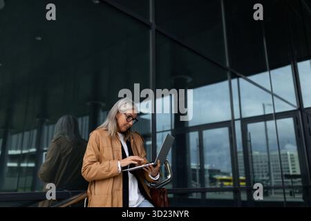 Élégante femme de cinquante ans travaillant sur un ordinateur portable à l'entrée de l'immeuble de bureaux Banque D'Images