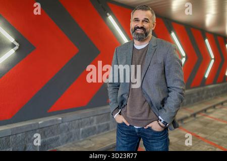 Portrait d'un homme d'affaires barbu mature avec un sourire dans une veste élégante grise contre un mur avec des flèches rouges Banque D'Images