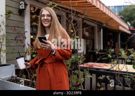 Une jeune femme avec une tasse de café dans les mains et un téléphone portable regarde au loin avec le sourire, attendant un jeune homme Banque D'Images