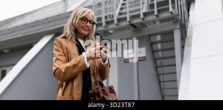 Femme d'affaires de cinquante ans avec un téléphone portable dans les mains dans un bâtiment industriel en toile de fond Banque D'Images