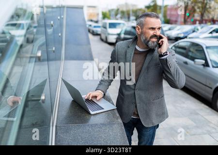 Homme d'affaires mature dans une veste avec un ordinateur portable parlant au téléphone à l'extérieur Banque D'Images