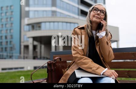 Une femme en âge de retraite avec un ordinateur portable parle sur un téléphone portable près du bureau Banque D'Images