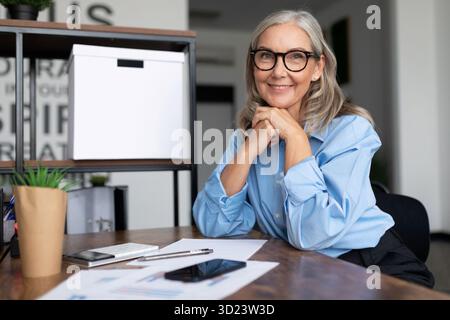 Avec succès la tête d'une femme d'affaires âgée d'âge moyen est assise à une table et regarde la caméra avec ses mains serrées dans une serrure, courir Banque D'Images