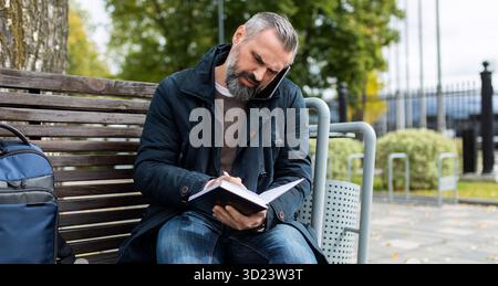 Femme d'affaires parlant au téléphone prendre des notes dans un cahier tout en étant assise sur un banc dans un parc de la ville Banque D'Images
