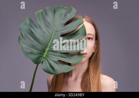 Jeune modèle féminin caucasien avec la feuille de monstera couvrant le visage sur fond gris Banque D'Images