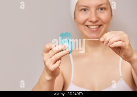 Femme caucasienne jeune adulte souriant tout en tenant du fil dentaire avec récipient bleu dans la salle de bain. Banque D'Images