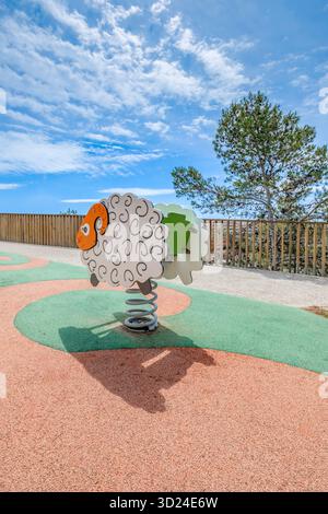 Cavalier de printemps de mouton vide sur une aire de jeux moderne avec plancher en caoutchouc, clôture en bois, et sapin par une journée ensoleillée Banque D'Images