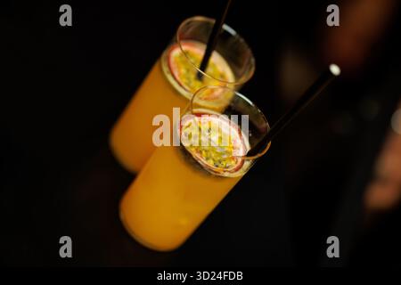 Deux grands verres de cocktails aux fruits de la passion avec glace et pailles noires servis sur fond sombre. Banque D'Images